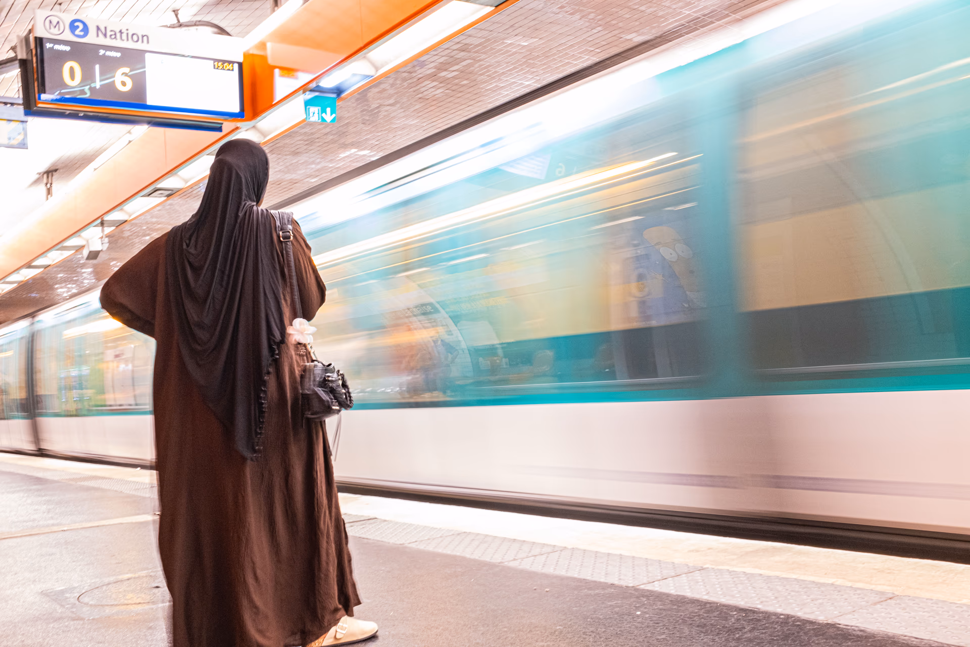 Frau in braunem Mantel wartet an U-Bahn-Station Nation in Paris, vorbeifahrender Zug mit Bewegungsunschärfe