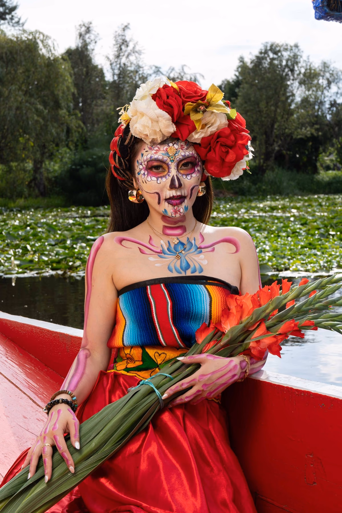 Mujer con maquillaje de Catrina y corona de flores sostiene gladiolas en una trajinera en el canal.