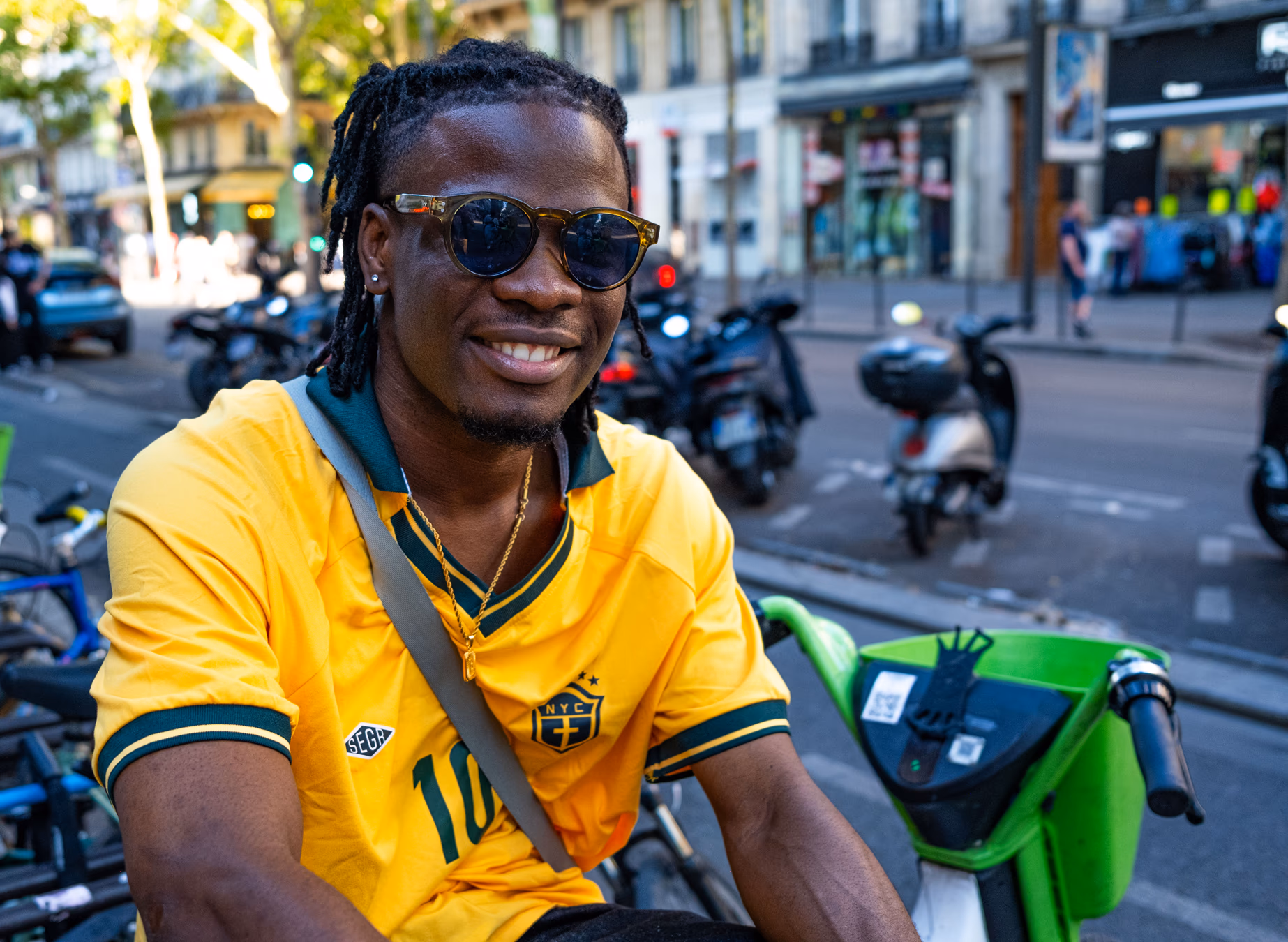 Lächelnder Mann in gelbem Shirt mit Sonnenbrille auf einer belebten Pariser Straße mit Motorrollern