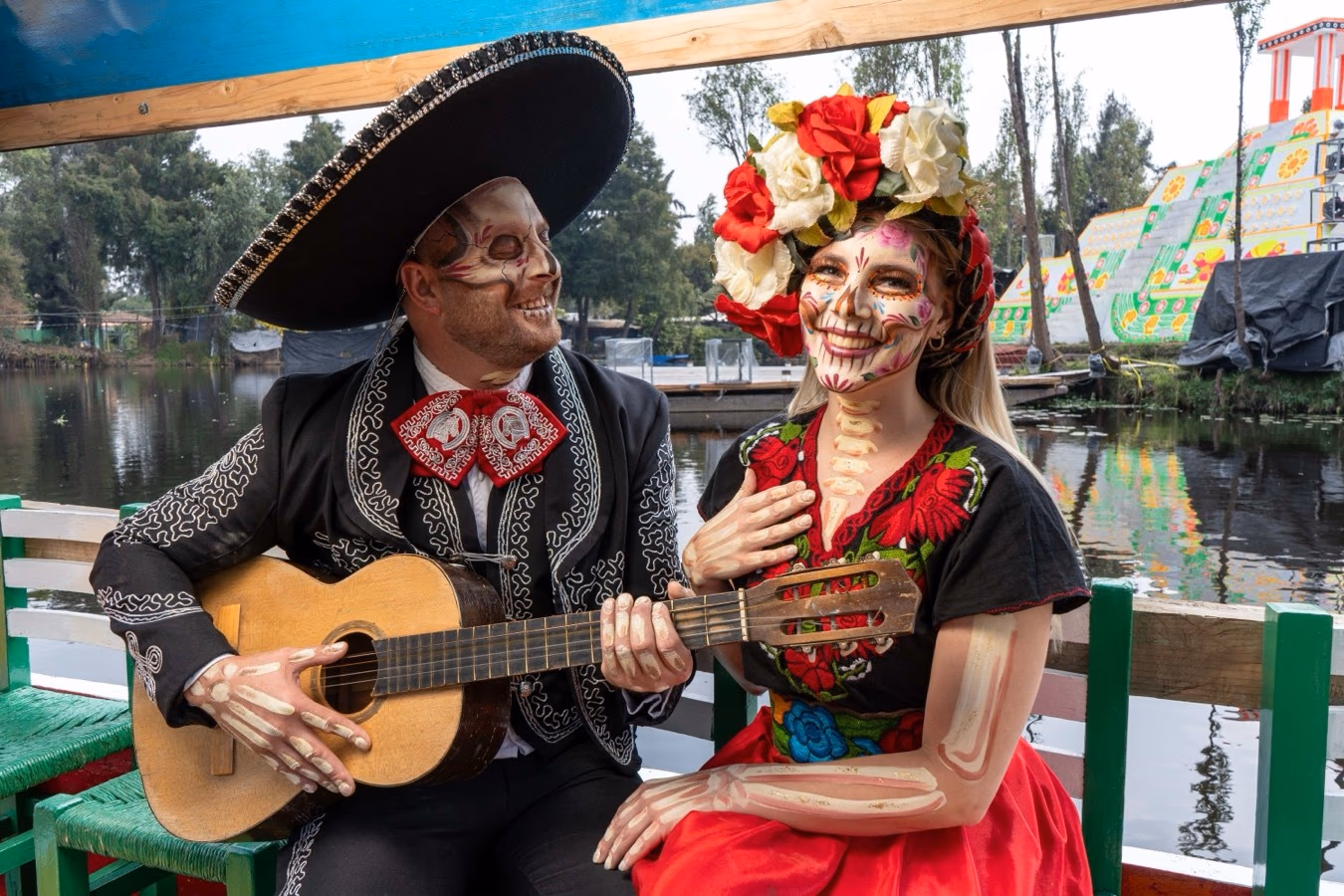 Hombre con guitarra y mujer con corona de flores, ambos maquillados como Catrinas, en una trajinera