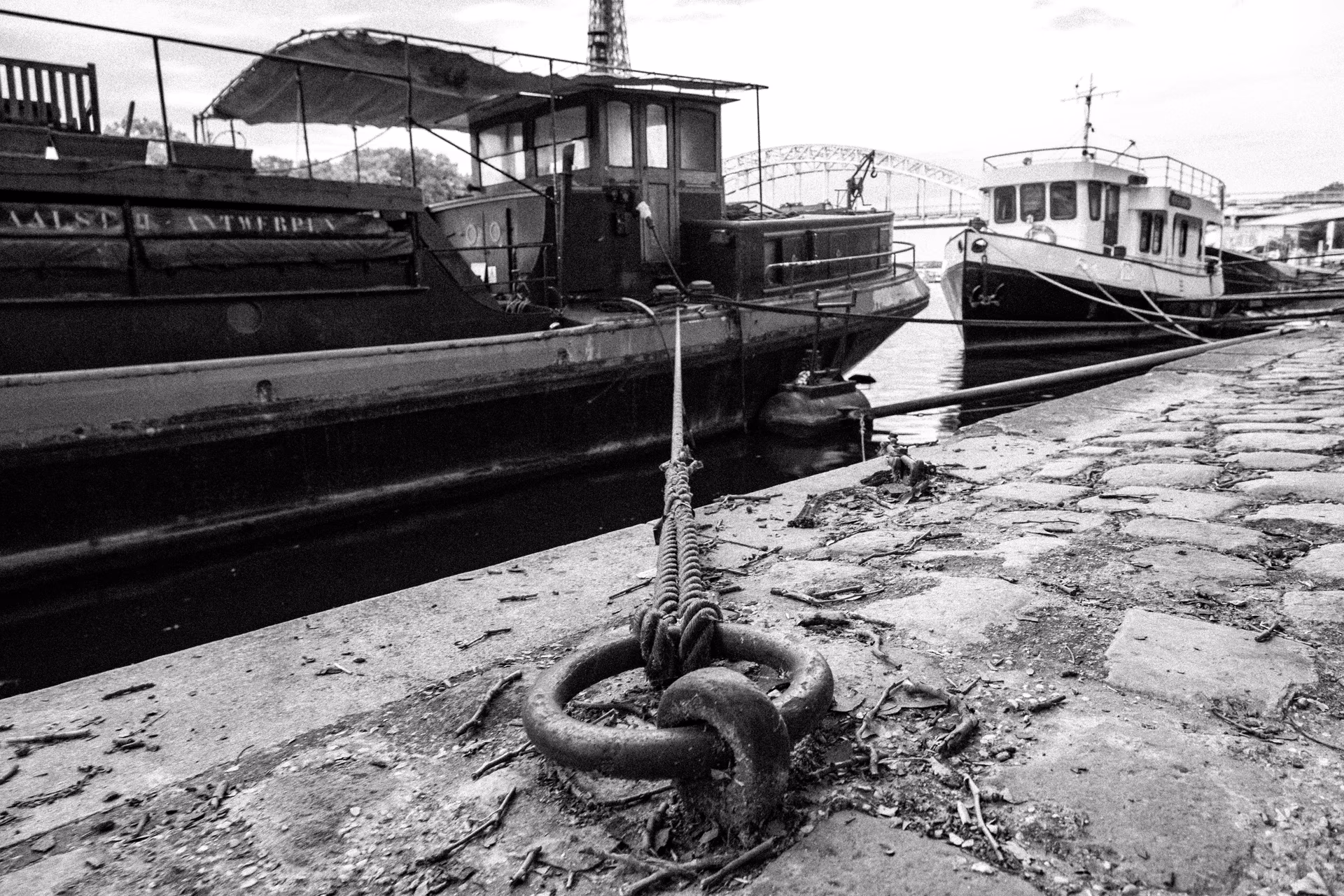 Schwarzweiß-Aufnahme von Flussbooten an einem Kai mit Ankerring im Vordergrund