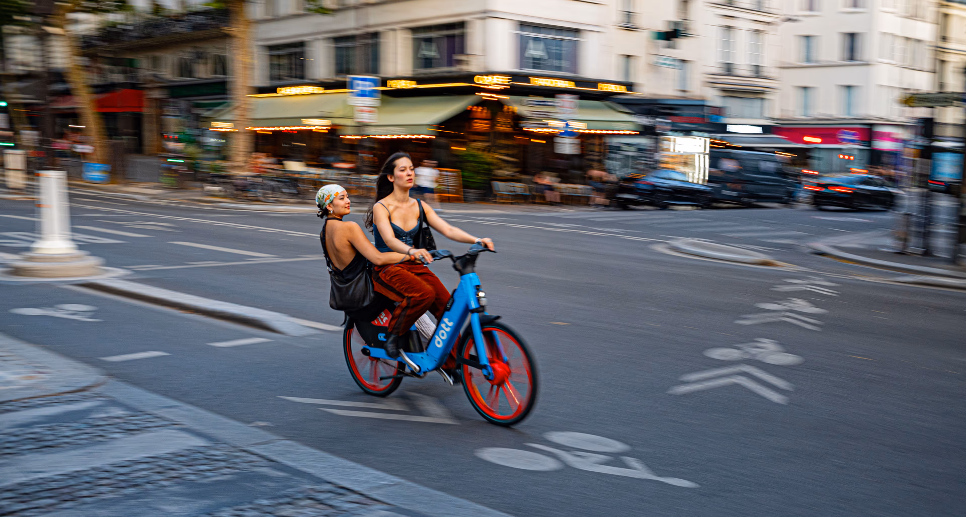 Zwei Frauen fahren auf einem roten und blauen Leihfahrrad durch eine belebte Pariser Straße.