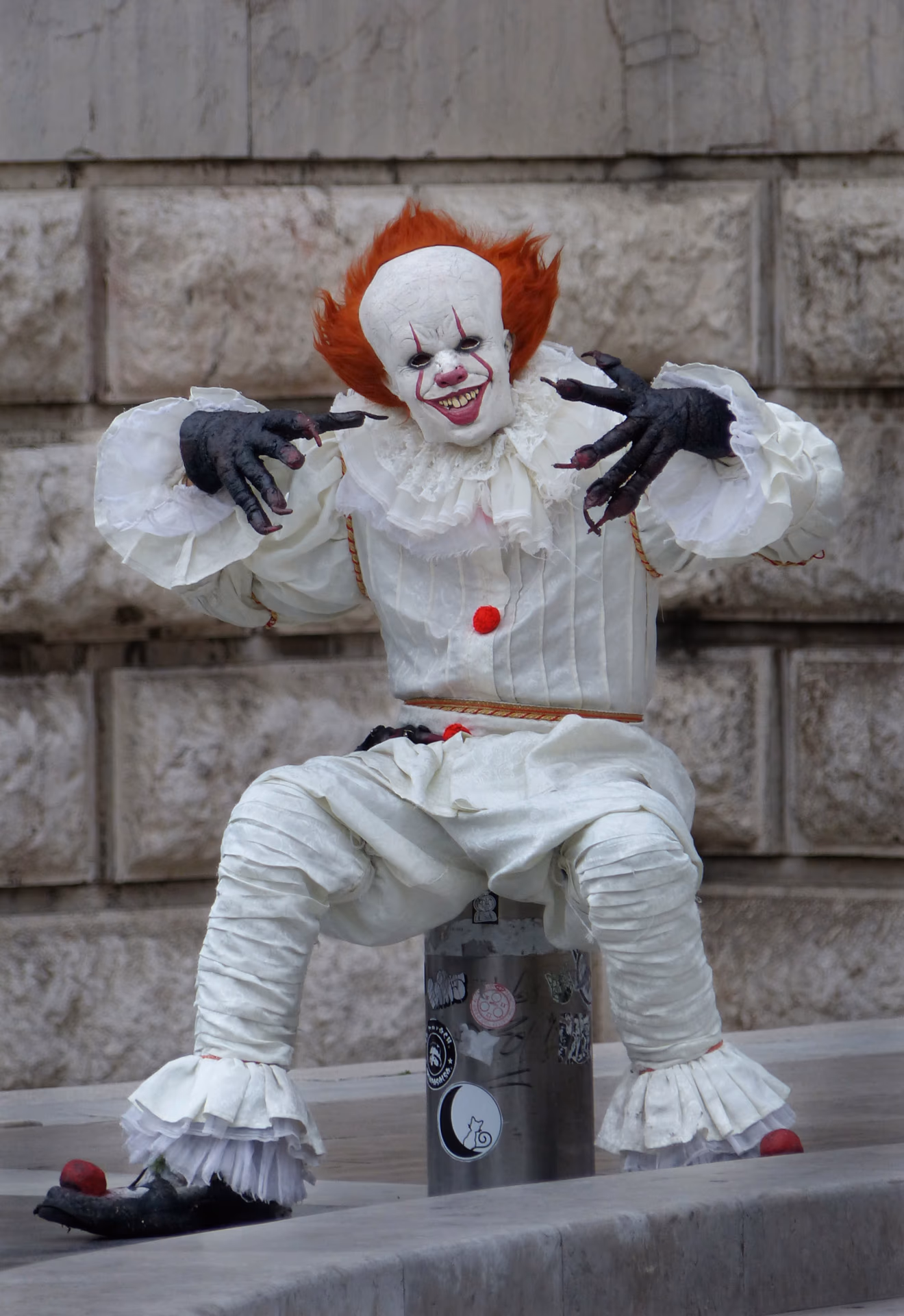 Gruseliger Clown mit roten Haaren und weißem Kostüm sitzt auf einer Säule.