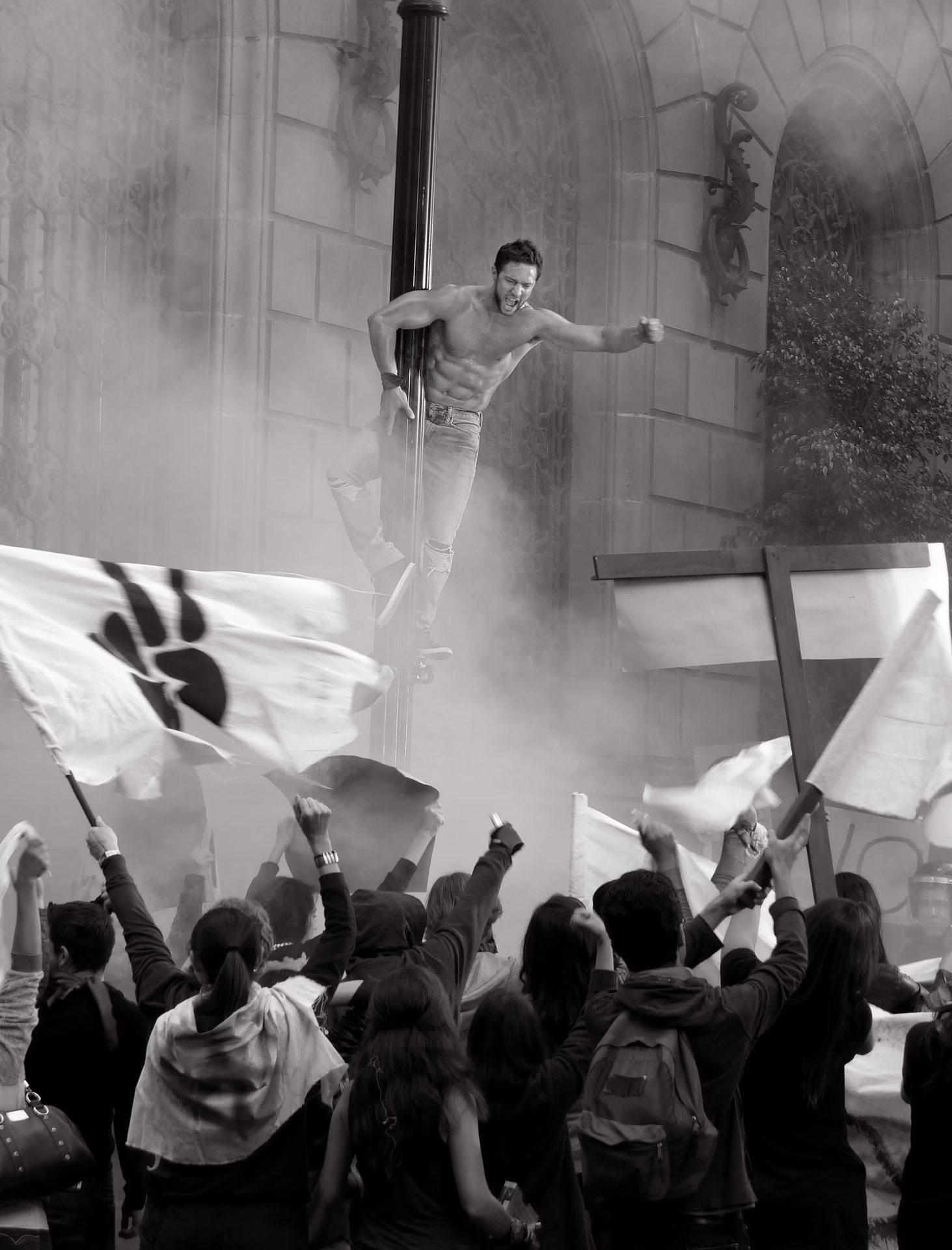 Mann mit nacktem Oberkörper klettert an einer Laterne, während eine Menge mit Fahnen protestiert.